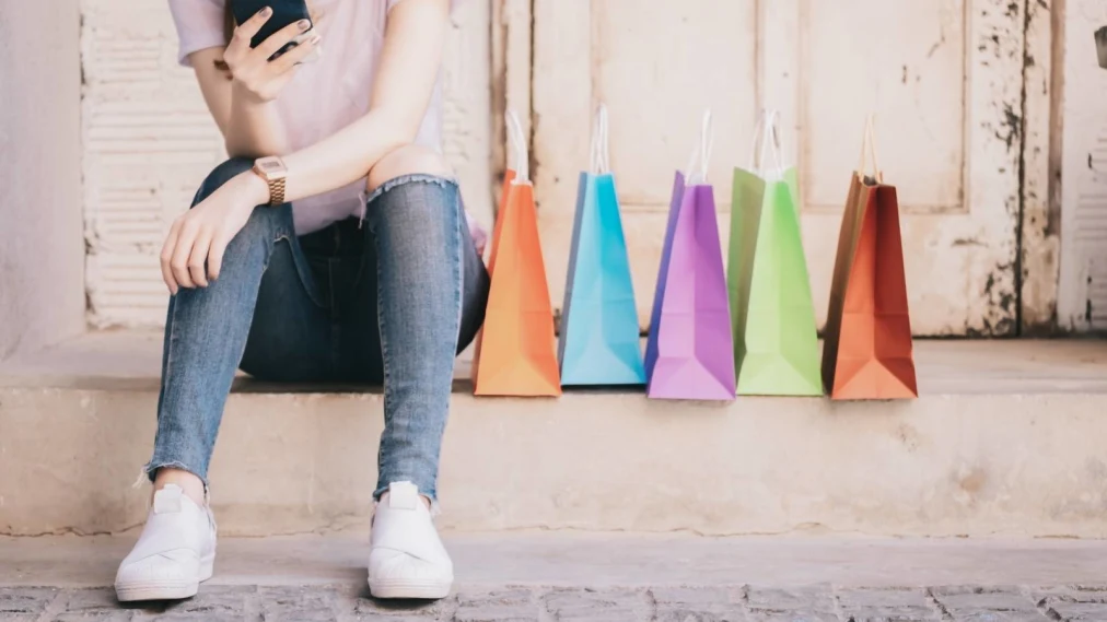 Person in blue jeans and tshirt sat on a step using a mobile phone with colourful shopping bags next to.