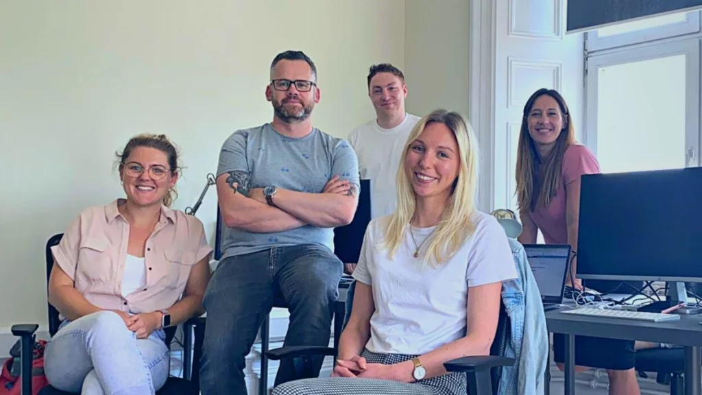 5 people sat in an office smiling at camera. Three women and 2 men.