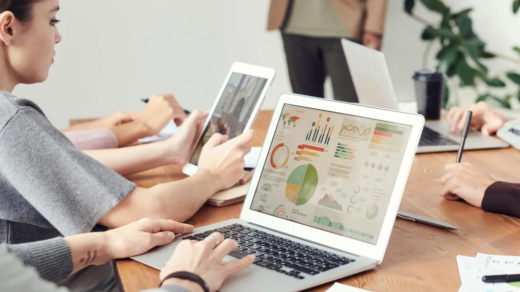 People around a desk working on ipads and laptops with images of graphs displayed.