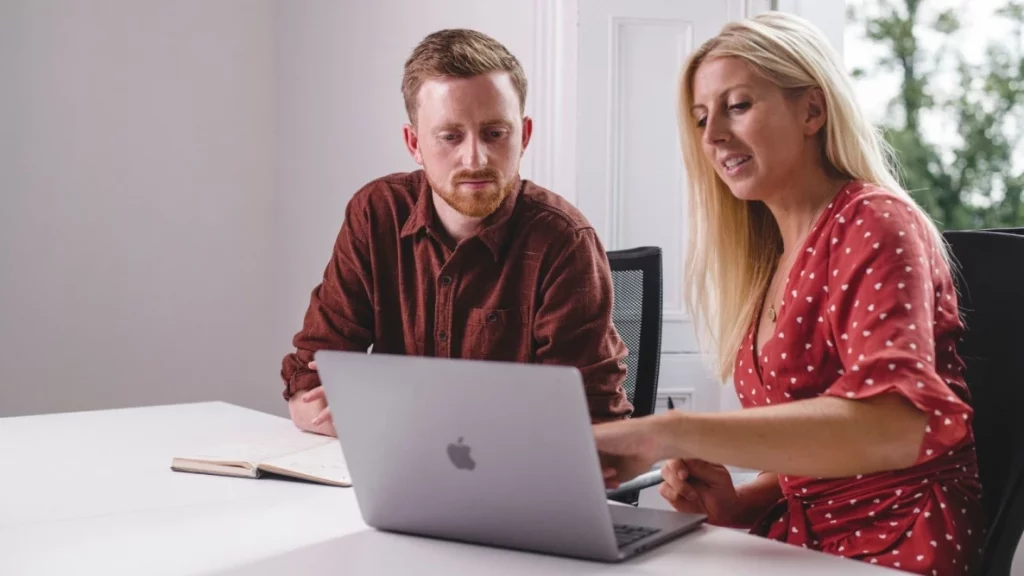 Man with beard and blonde haired lady sat at table looking at a laptop screen. Lady is pointing at screen, man is making notes.