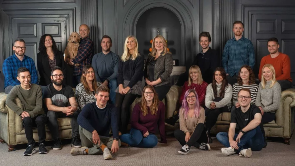 The Launch Online Team looking smart. All looking at the camera smiling. Some stood and some sat on the floor and sofa.
