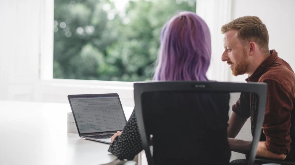 Back of two peoples heads - man and woman, sat at a table and looking at Google Analytics together on a laptop screen.