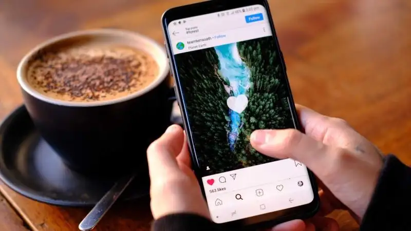 Hands holiding a mobile phone scrolling Instagram with a coffee in the background