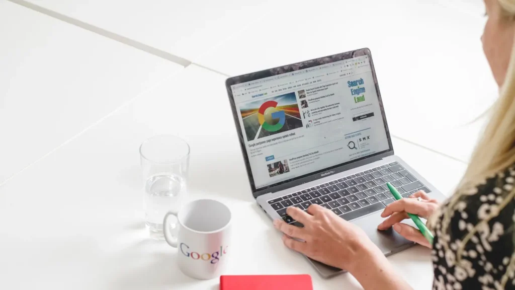 Becky - blonde woman - sat at a table using a laptop looking at Google News with a Google mug and glass of water