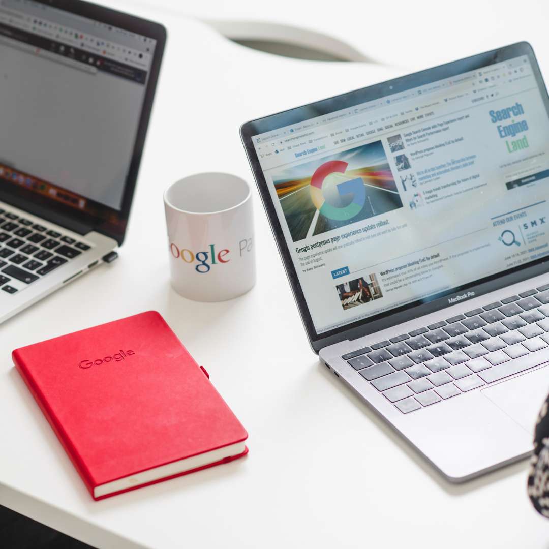 Desk with a Google mug, red notebook and laptops showing Google on the screens