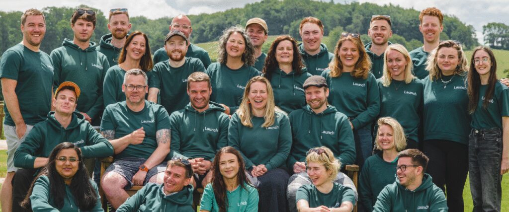 The performance marketing team at Launch stands and smiles in front of rolling green hills.