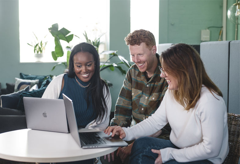 Image shows team of three people sitting around a laptop chatting and smiling.